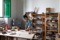 Senhora adulta focada em roupas casuais e avental esculpir peça de argila e sorrir durante a aula de cerâmica na oficina — Fotografia de Stock