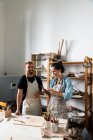 Mujer de mediana edad positiva y hombre joven en ropa casual y delantales sonriendo mientras crea placa de arcilla durante la clase de cerámica en el taller - foto de stock