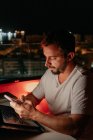 Freelancer masculino jovem concentrado em mensagens de roupa casual no smartphone enquanto se senta no telhado do edifício moderno e trabalha remotamente no laptop à noite — Fotografia de Stock