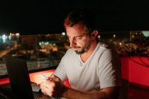 Freelancer masculino jovem concentrado em mensagens de roupa casual no smartphone enquanto se senta no telhado do edifício moderno e trabalha remotamente no laptop à noite — Fotografia de Stock