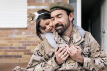 Cute daughter tenderly embracing father in military uniform sitting at doorstep after arrival — Stock Photo