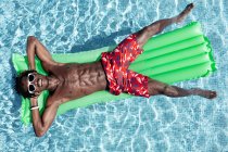 From above of carefree African American male with naked torso in shorts and sunglasses lying on inflatable mattress in swimming pool and enjoying sunny day during summer holiday — Stock Photo