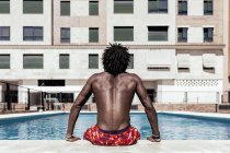 Vue arrière d'un homme afro-américain torse nu et méconnaissable assis au bord de la piscine et profitant d'une journée ensoleillée pendant les vacances d'été — Photo de stock