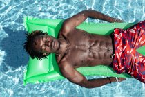 From above of carefree African American male with naked torso and in shorts lying eyes closed on inflatable mattress in swimming pool and enjoying sunny day during summer holiday — Stock Photo