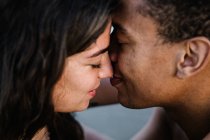 Viajero masculino afroamericano besando a su pareja femenina sincera de pie contra el océano durante el viaje de verano - foto de stock