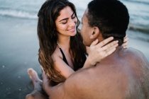 Young content barefoot multiracial couple embracing on sandy ocean beach during summer trip looking at each other — Stock Photo