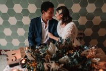 Jovem noiva e noivo multirracial sentados à mesa com buquê de flores e velas flamejantes enquanto olham um para o outro no restaurante — Fotografia de Stock