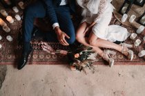 De arriba recortado irreconocible pareja de recién casados jóvenes con ropa de boda elegante sentado en el suelo en la alfombra árabe - foto de stock