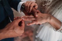 Высокоугольный вид сбоку обрезанный неузнаваемый этнический жених надевает кольцо на палец невесты в модных свадебных платьях, нежно держась за руки с любовью — стоковое фото