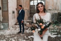 Content young bride wearing classy wedding gown with tender bouquet standing looking at camera near ethnic groom in obsolete ruined building room — Stock Photo