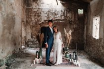 Side view full body loving young ethnic newlywed couple wearing chic wedding clothes embracing and kissing gently in obsolete remaining building — Stock Photo