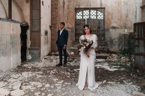Contenu jeune mariée portant une robe de mariée chic avec un bouquet tendre debout regardant la caméra près du marié ethnique dans une pièce de bâtiment désuète en ruine — Photo de stock