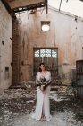 Ganzkörperheitere junge ethnische Braut in elegantem weißen Kleid mit zartem Strauß steht in verlassenen Ruinen und blickt in die Kamera — Stockfoto
