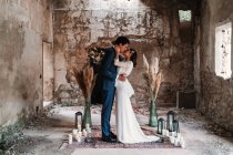 Seitenansicht Ganzkörperliebe junge ethnische Brautpaar trägt schicke Hochzeitskleidung umarmen und küssen sanft in veralteten verbleibenden Gebäude — Stockfoto