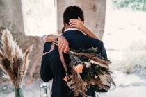 Наречений на задньому плані в елегантному тукседо обіймає наречену з букетом в гранжевому зовнішньому будівництві в сонячний день — стокове фото