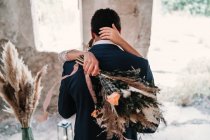 Back view groom in elegant tuxedo hugging bride with bouquet in grunge outdoor construction on sunny day — Foto stock