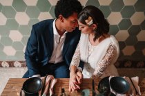 Von oben streicheln sich junge multiethnische Braut und Bräutigam, während sie mit Blumenstrauß und brennenden Kerzen im Restaurant mit geschlossenen Augen am Tisch sitzen — Stockfoto