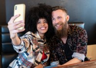 Захоплена багаторасова пара сидить за столом в кафе і приймає селфі на смартфон — стокове фото