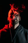 Serious male rocker smoking cigarette and looking at camera on black background in studio with red illumination — Stock Photo
