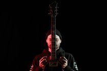 Tranquilo músico de rock masculino de pie con guitarra eléctrica sobre fondo negro en estudio con luz roja - foto de stock