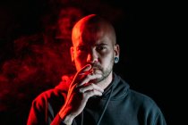 Серьезный мужчина рокер курить сигарету и смотреть на камеру на черном фоне в студии с красным освещением — стоковое фото
