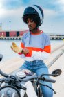 Concentrated young black female biker with Afro hair in trendy outfit and helmet wearing gloves while sitting on motorcycle at seaside — Stock Photo