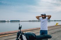 Thoughtful young ethnic unshaven male in casual outfit and cap standing near modern motorcycle with hands behind head and looking away near port — Stock Photo