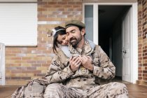 Cute daughter tenderly embracing father in military uniform sitting at doorstep after arrival — Stock Photo