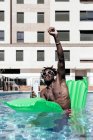 Delighted African American male sitting on inflatable mattress in swimming pool and listening to music in headphones while enjoying summer vacation with raised arm — Stock Photo