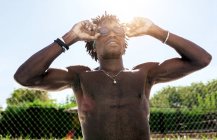 Joven atleta afroamericano confiado con torso desnudo ajustando gafas y mirando hacia arriba mientras está de pie junto a la piscina después de nadar - foto de stock