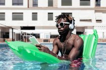 Восхитительный афроамериканец, сидящий на надувном матрасе в бассейне и слушающий музыку в наушниках, наслаждаясь летними каникулами — стоковое фото