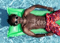 From above of carefree African American male with naked torso in shorts and sunglasses lying on inflatable mattress in swimming pool and enjoying sunny day during summer holiday — Stock Photo