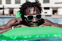 Hombre afroamericano fresco en gafas de sol acostado en un colchón inflable y disfrutando del fin de semana en la piscina - foto de stock