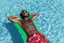 Desde arriba de despreocupado hombre afroamericano con torso desnudo en pantalones cortos y gafas de sol tumbados en colchón inflable en la piscina y disfrutando de un día soleado durante las vacaciones de verano - foto de stock