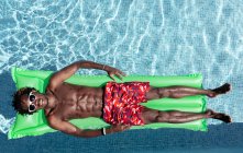 Von oben ein unbeschwerter afroamerikanischer Mann mit nacktem Oberkörper in kurzen Hosen und Sonnenbrille, der auf aufblasbarer Matratze im Schwimmbad liegt und den sonnigen Tag im Sommerurlaub genießt — Stockfoto