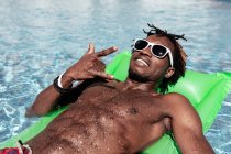 Hombre afroamericano fresco en gafas de sol tumbado en un colchón inflable y mostrando un gesto de roca mientras mira a la cámara y disfruta del fin de semana en la piscina - foto de stock