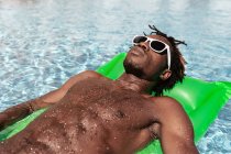 Охолоджений афро-американець у сонцезахисних окулярах лежить на надувному матраці і насолоджується вихідними в басейні — стокове фото
