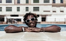 Deleitado hombre afroamericano en gafas de sol apoyadas en la piscina mientras disfruta de las vacaciones de verano y mira a la cámara - foto de stock