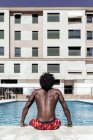Vista trasera de un irreconocible hombre afroamericano sin camisa sentado junto a la piscina y disfrutando de un día soleado durante las vacaciones de verano - foto de stock