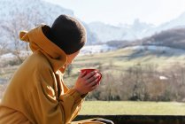 Side view contemplative female in warm clothes and hood holding cup of aromatic hot drink and admiring scenic highlands on sunny winter day — Stock Photo