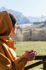 Side view contemplative female in warm clothes and hood holding cup of aromatic hot drink and admiring scenic highlands on sunny winter day — Stock Photo