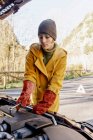 Junge Frau in warmer Kleidung und nicht leitenden Handschuhen klemmt Autobatterie unter Motorhaube im sonnigen Herbstpark — Stockfoto