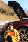 Calm young female in warm clothes having phone call and sitting on the floor leaning on car with opened hood on roadside and looking away after breakdown on sunny autumn day — Stock Photo