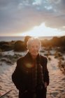 Felice turista anziana con i capelli grigi in abito casual caldo sorridente e guardando la fotocamera mentre si rilassa sulla spiaggia sabbiosa contro cielo nuvoloso sera — Foto stock