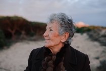 Happy elderly female tourist with gray hair in warm casual outfit smiling and looking away while relaxing on sandy beach against cloudy evening sky — Stock Photo