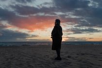 Seitenansicht einer nicht wiedererkennbaren Seniorin in warmer Freizeitkleidung, die den Sonnenuntergang über dem Meer bewundert, während sie sich allein am Sandstrand ausruht — Stockfoto