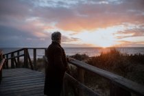 Side view of anonymous elderly female traveler in casual clothes standing on wooden pier on sandy beach and enjoying seascape at sunset — Stock Photo