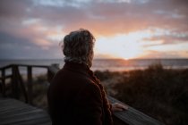 Side view of elderly female traveler in casual clothes standing on wooden pier on sandy beach and enjoying seascape at sunset — Stock Photo