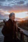 Seitenansicht einer älteren Reisenden in Freizeitkleidung, die auf einem hölzernen Pier am Sandstrand steht und das Meer bei Sonnenuntergang genießt — Stockfoto