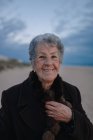 Feliz turista anciana con el pelo gris en traje casual cálido sonriendo y mirando a la cámara mientras se relaja en la playa de arena contra el cielo nublado de la noche - foto de stock